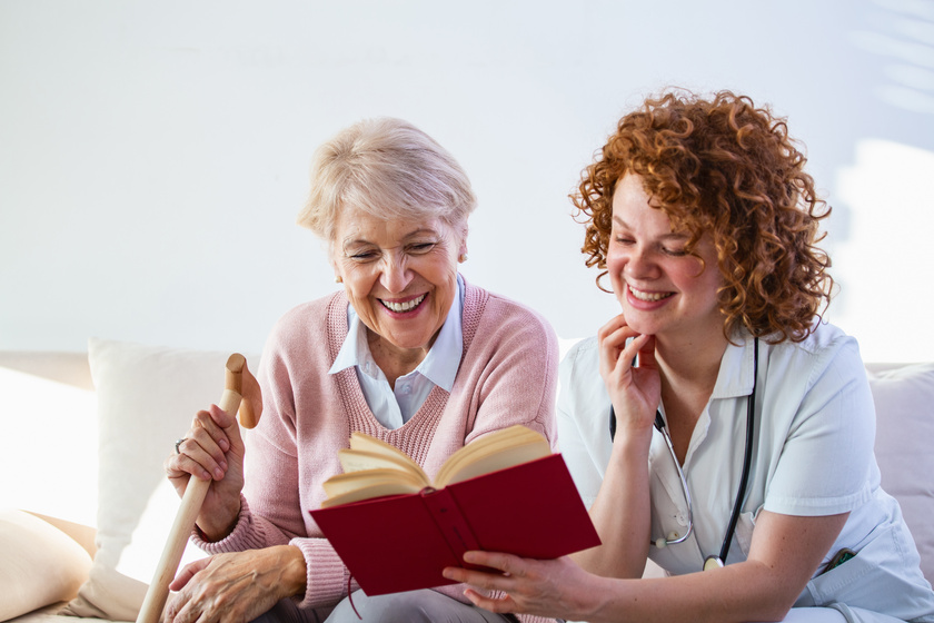 Woman caregiver reading a book while sitting with happy senior woman at nursing home. Happy elder woman sitting on white sofa and listening to nurse reading a book out loud Woman caregiver reading a book while sitting with happy senior woman at nursing home. Happy elder woman sitting on white sofa and listening to nurse reading a book out loud