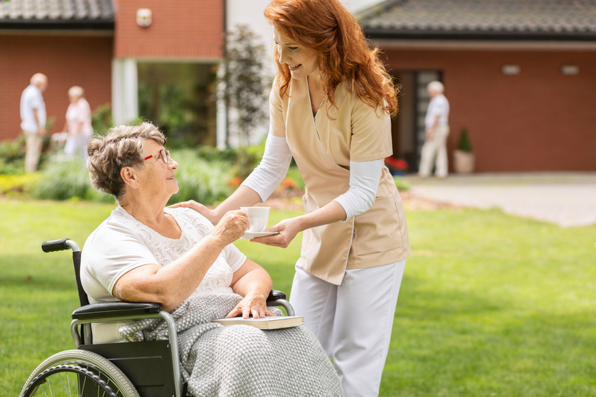 Friendly nurse giving tea to disabled senior woman in the wheelc