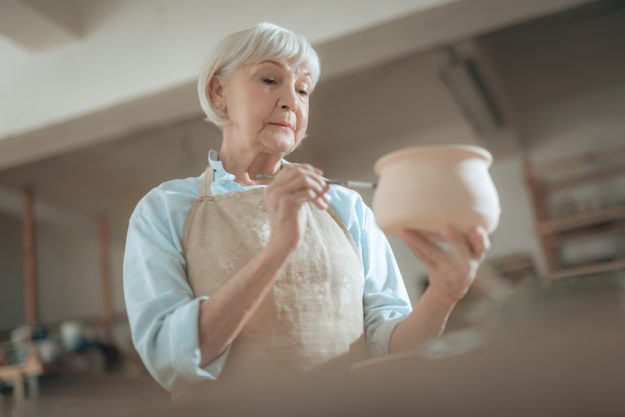 Low angle of old craftswoman painting her pot in workshop Why Pottery Is A Good Activity When Staying At Active Adult Homes