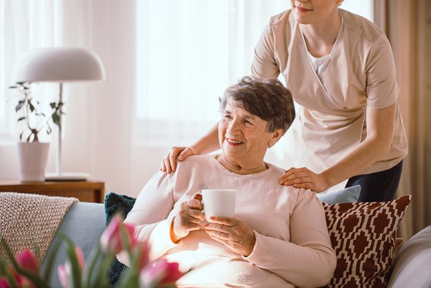 Smiling senior woman drinking tea with her caregiver standing behind her Smiling senior woman drinking tea with her caregiver standing behind her
