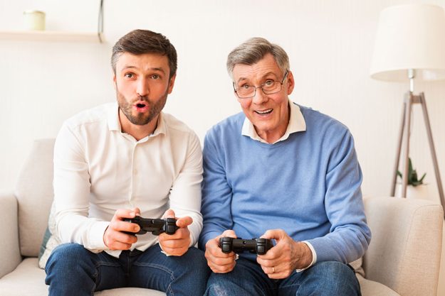 Son And Elderly Father Playing Videogame Sitting On Sofa