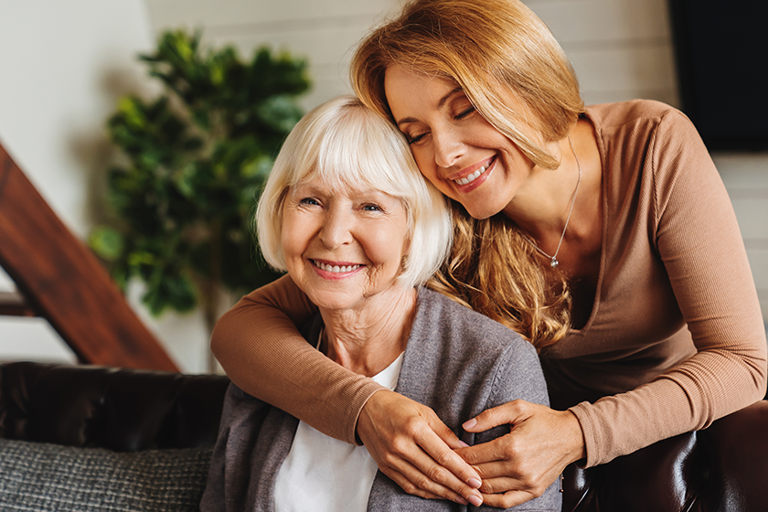 Happy daughter embracing from behind elderly mother at living room