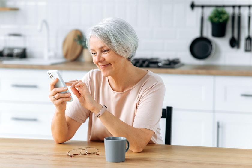 Happy pretty caucasian middle aged gray haired woman, sits at home in the kitchen, holds her smartphone in her hand, uses it to communicate in social networks, read news, makes an online order, smiles 5 Social Media Tips For Senior