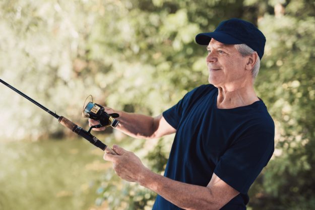 An elderly man stands on the river bank with a spinning in his hands, turns the fishing reel and stares at the water Exploring The Best Fishing Spots Near Your Senior Home In Fort Smith, AR