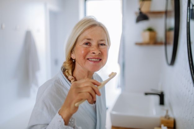 Beautiful senior woman in bathrobe brushing teeth with eco wooden toothbrush inbathroom, sustainable lifestyle. Beautiful senior woman in bathrobe brushing teeth with eco wooden toothbrush inbathroom, sustainable lifestyle.