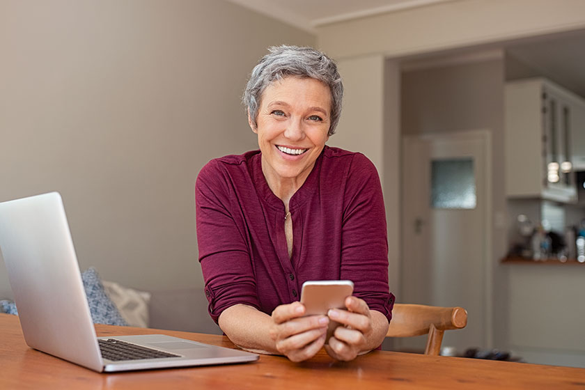 smiling casual senior woman using laptop while messaging smartphone
