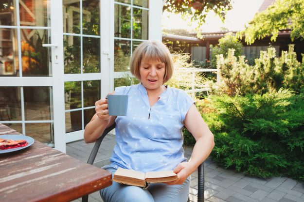 Middle aged woman reading book during breakfast in yard The Reasons Why Minimalism Is A Perfect Lifestyle For Those In Active Senior Living Apartments In Katy, TX