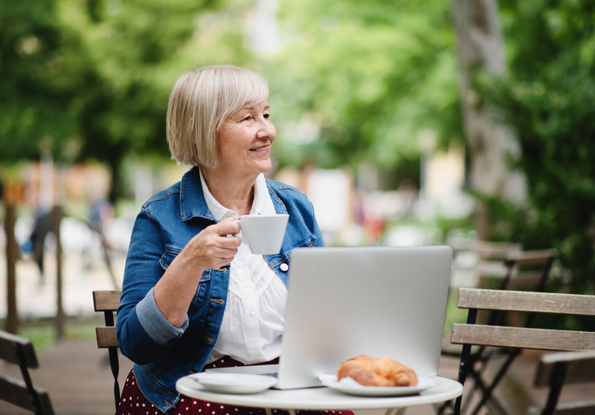 Senior woman with coffee sitting outdoors in cafe, using laptop.