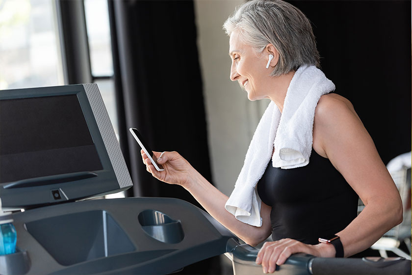 Happy retired woman in wireless earphone holding smartphone while standing treadmill