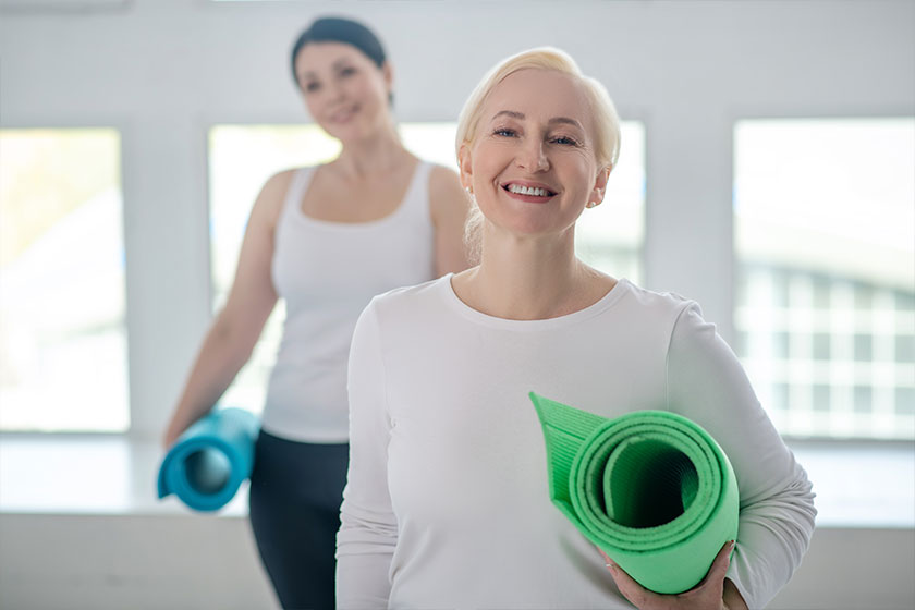 Blonde female standing with yoga rug and smiling brunette female standing behind her