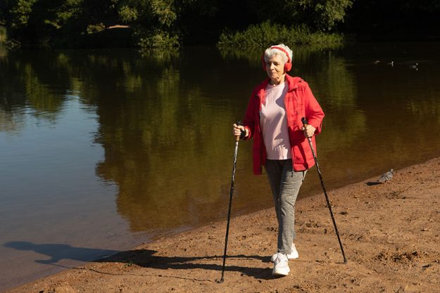 grey haired woman wearing red jacket walking tracking sticks beach