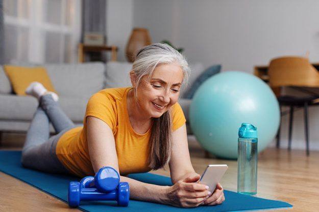 senior woman resting yoga mat browsing internet smartphone home while