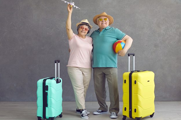 studio portrait of happy senior couple in hats and sunglasses with paper plane and suitcases