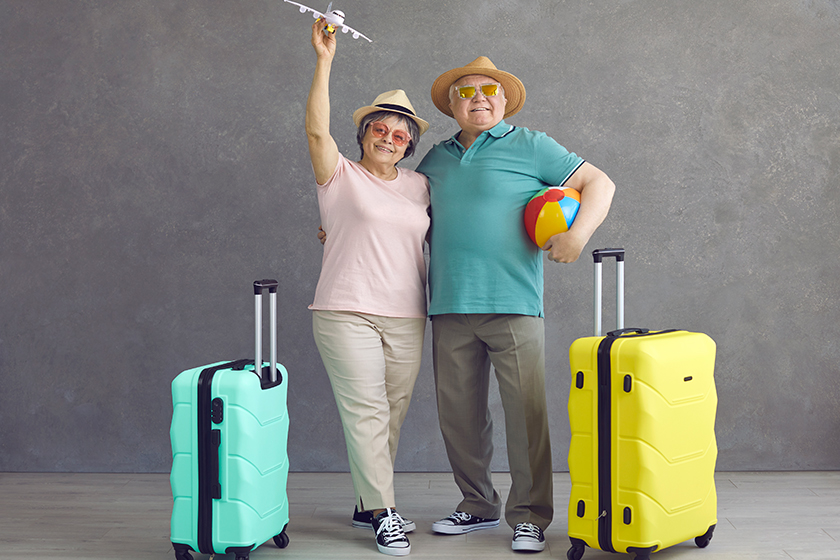 studio portrait of happy senior couple in hats and sunglasses with paper plane and suitcases
