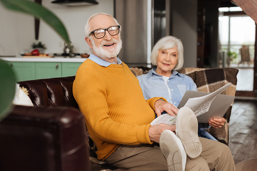 happy elderly husband newspaper looking camera wife couch blurred plant happy elderly husband newspaper looking camera wife couch blurred plant