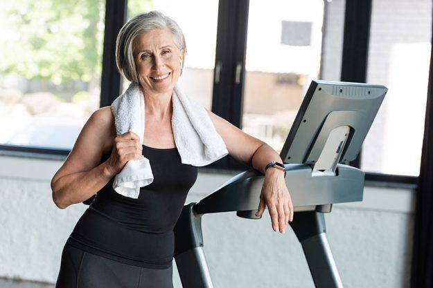 joyful retired woman white towel shoulders standing next treadmill