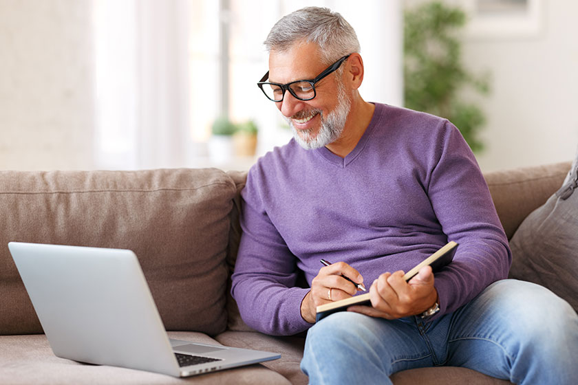 Handsome happy senior man in glasses working remotely while sitting on sofa with laptop computer
