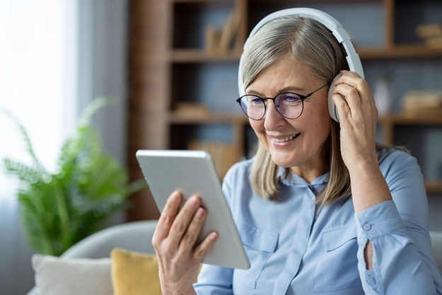 Happy senior woman wearing headphones using digital tablet while sitting Happy senior woman wearing headphones using digital tablet while sitting