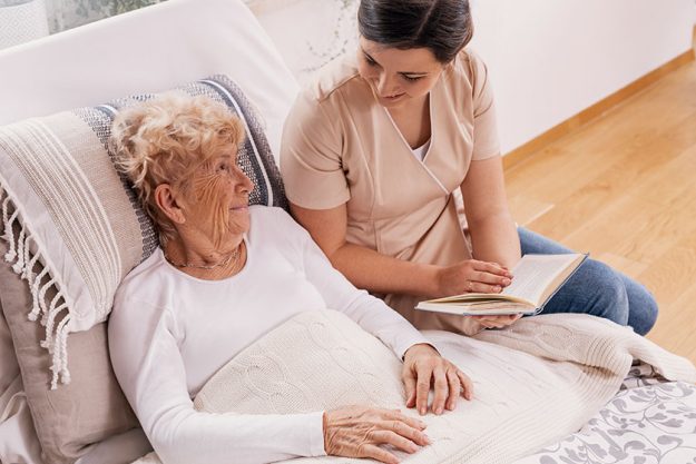 Helpful female volunteer reads book elderly woman lying bed