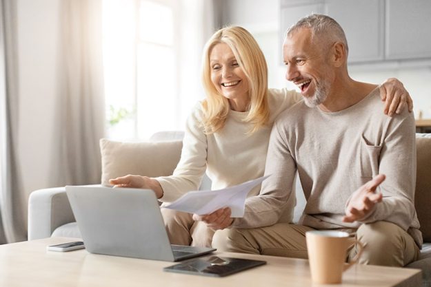 Smiling mature couple discussing happily papers laptop sitting sofa bright