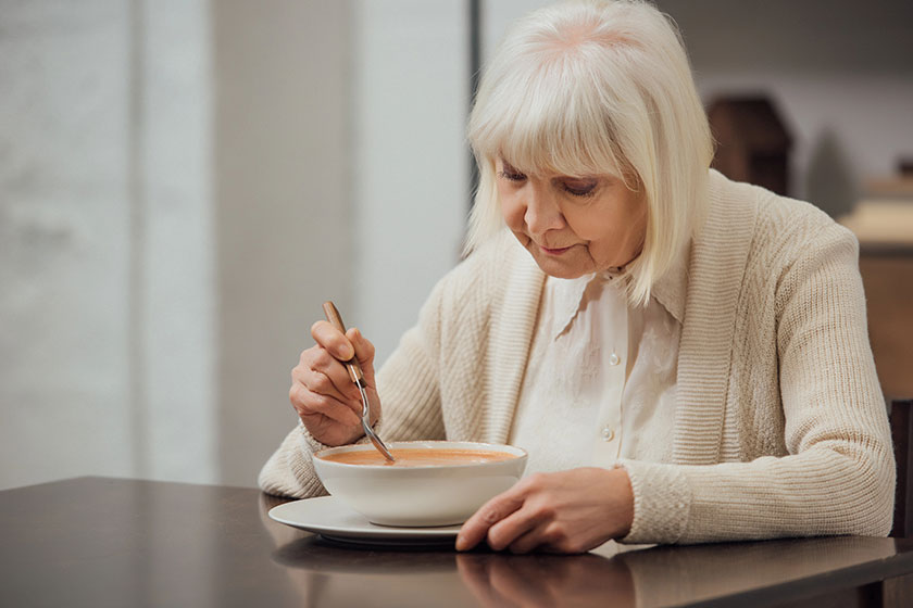 Senior woman grey hair sitting table eating cream soup home
