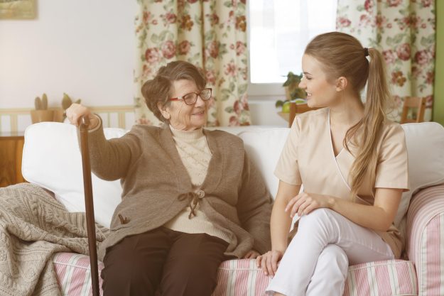 Young,Caregiver,And,Senior,Woman,Sitting,On,A,Sofa,In Young,Caregiver,And,Senior,Woman,Sitting,On,A,Sofa,In