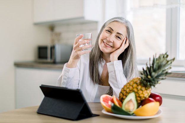 Beautiful retired gray haired woman with glass of water in kitchen sitting at the table Beautiful retired gray haired woman with glass of water in kitchen sitting at the table