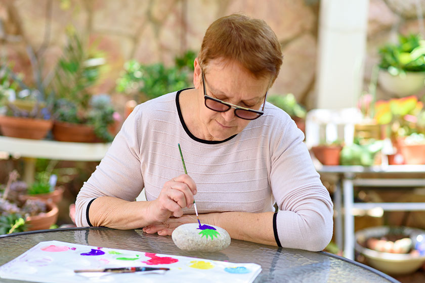 Happy authentic senior woman sitting outdoors table painting pictures stone Happy authentic senior woman sitting outdoors table painting pictures stone