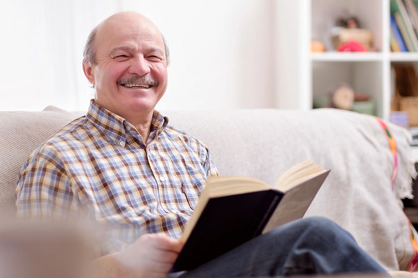 Senior man reading book smiling looking camera sitting his sofa Senior man reading book smiling looking camera sitting his sofa