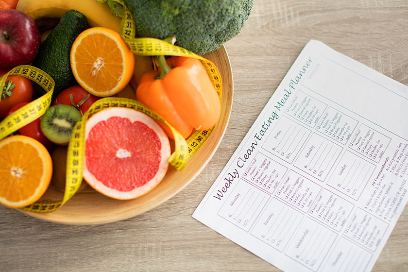 Weekly eating meal planner and plate of fresh fruits and vegetables nearby on table Weekly eating meal planner and plate of fresh fruits and vegetables nearby on table