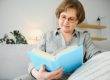Restful reading. Portrait of thoughtful aged woman reading favorite literature at cozy home. She is lying on pillows on comfortable sofa. Legs are covered with soft blanket.