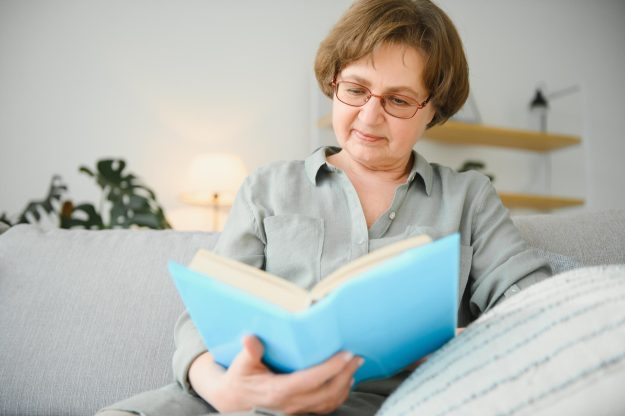 Restful reading. Portrait of thoughtful aged woman reading favorite literature at cozy home. She is lying on pillows on comfortable sofa. Legs are covered with soft blanket.