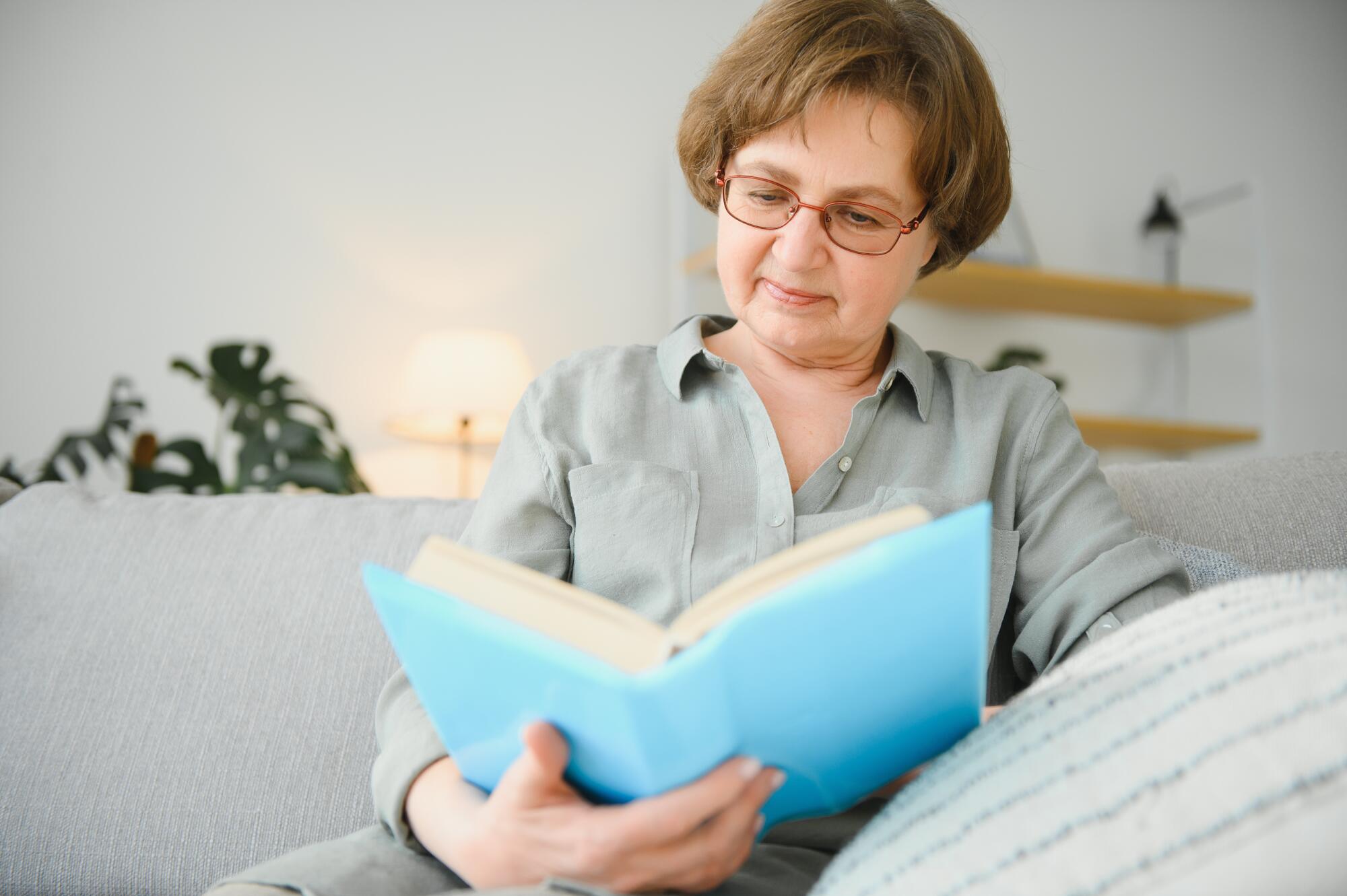 Restful reading. Portrait of thoughtful aged woman reading favorite literature at cozy home. She is lying on pillows on comfortable sofa. Legs are covered with soft blanket. Restful reading. Portrait of thoughtful aged woman reading favorite literature at cozy home. She is lying on pillows on comfortable sofa. Legs are covered with soft blanket.