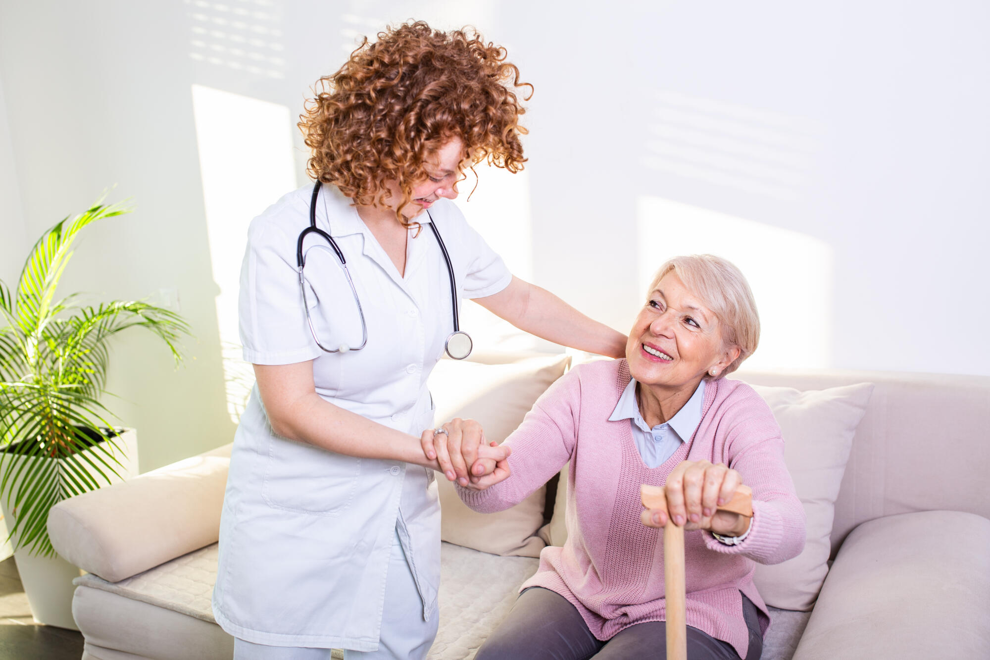 Female caregiver helping senior woman get up from couch in living room. Smiling nurse assisting senior woman to get up. Caring nurse supporting patient while getting up from sofa Female caregiver helping senior woman get up from couch in living room. Smiling nurse assisting senior woman to get up. Caring nurse supporting patient while getting up from sofa